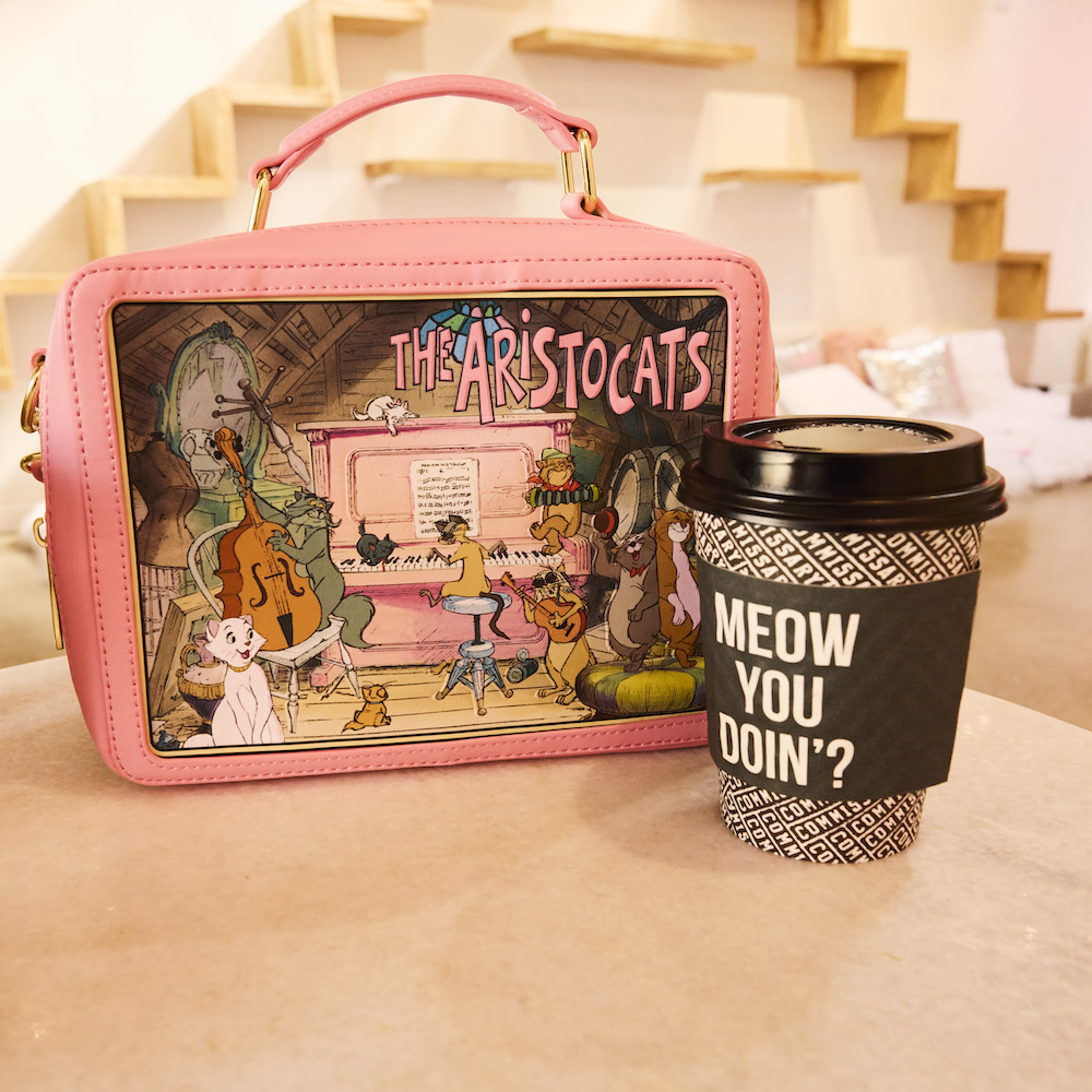 Pink crossbody bag in the shape of a lunchbox featuring The Aristocats next to a cup of coffee.